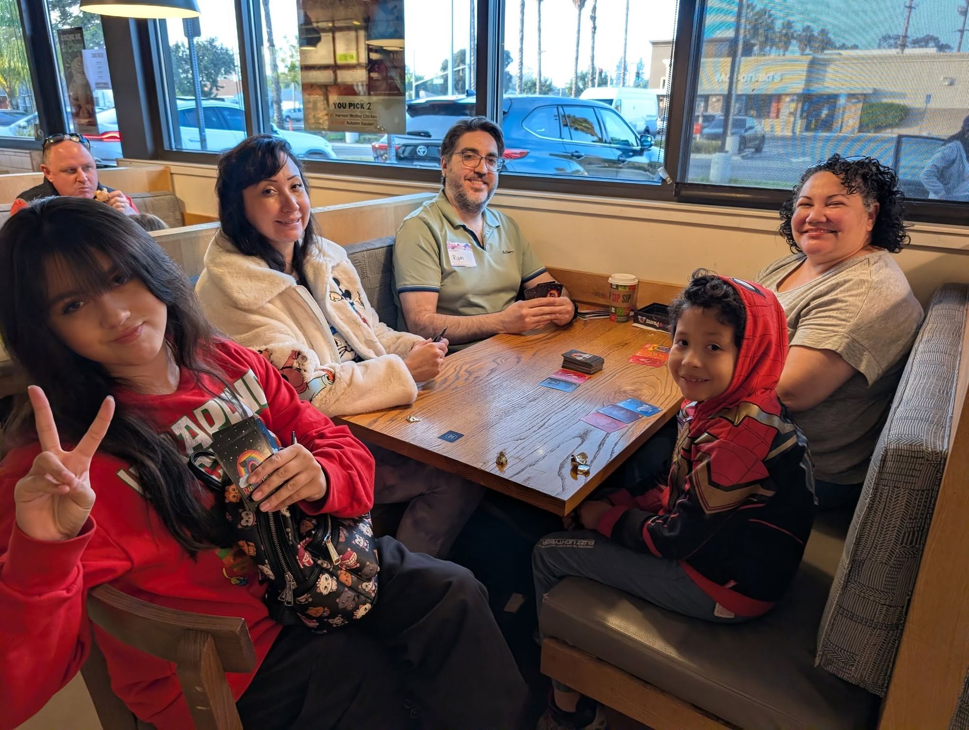 Multigenerational Rummy Club group at a table, including a young woman giving a peace sign, a man with a name tag, and a kid in a Spider-Man hoodie
